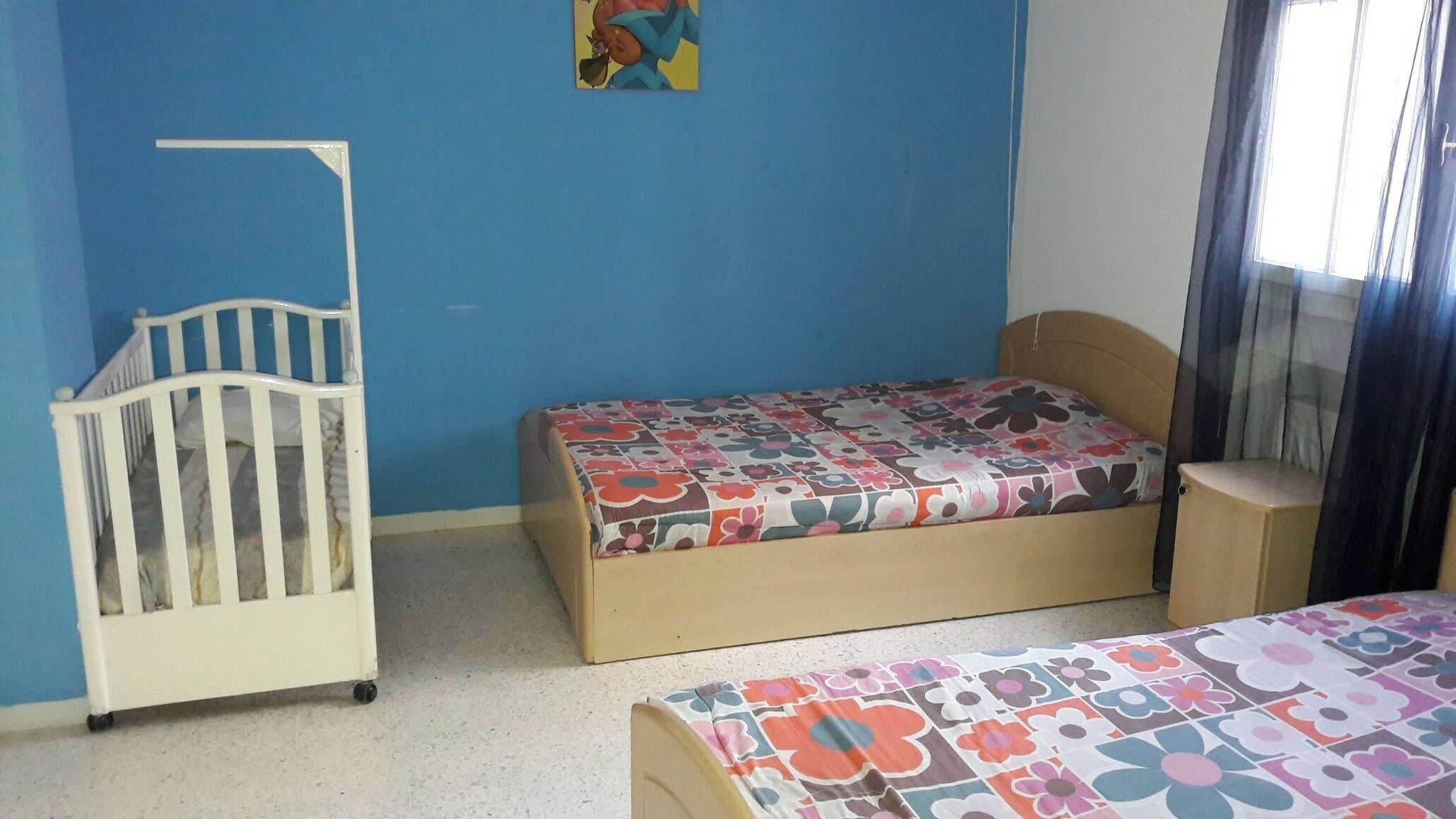 A room with a crib, a bed with a floral bedspread, and a window with curtains.