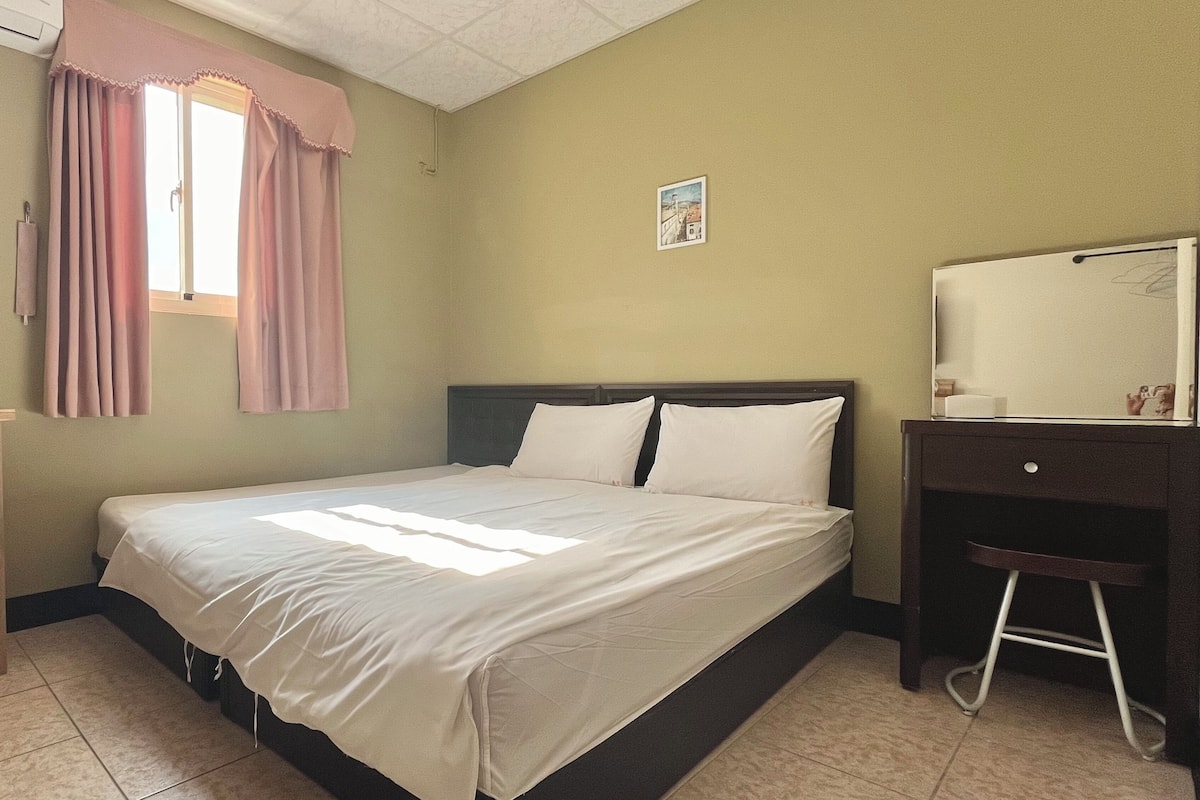 Habitación de hotel con cama, escritorio, silla, ventana con cortinas y televisor montado en la pared.