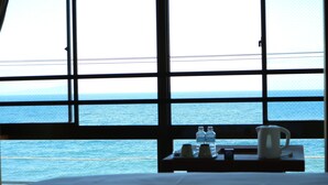 A room with a large window overlooking the sea, a bed, a small table with a kettle, and two bottles. - Hotel & Resort Izunone (Higashiizu)