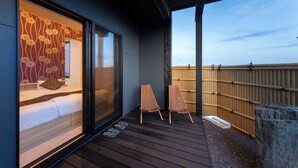 Une pièce comprenant un lit, une porte-fenêtre coulissante, deux chaises en bois et donnant sur une clôture en bambou.