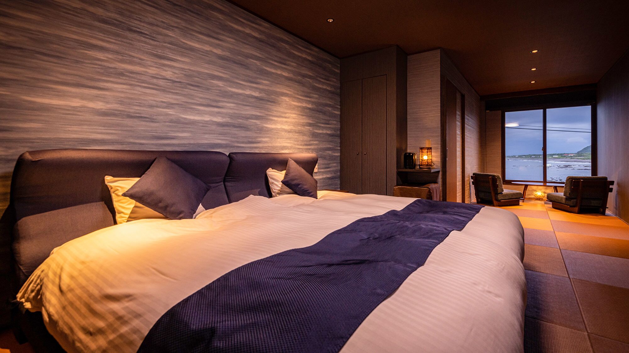 A large bed with a headboard, a nightstand with a lamp, and a view of the sea.
