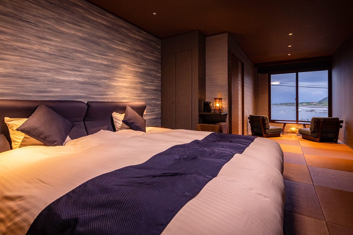Een groot bed met hoofdeinde, een nachtkastje met lamp en uitzicht op zee.