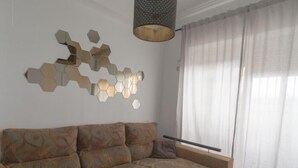 Un salón con sofá modular, una mesa de centro de vidrio, un aplique hexagonal empotrado en la pared, una lámpara colgante y cortinas traslúcidas.
