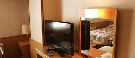 A hotel room with a desk, a television, a mirror, a refrigerator, and a bedside table with a lamp and a tissue box.