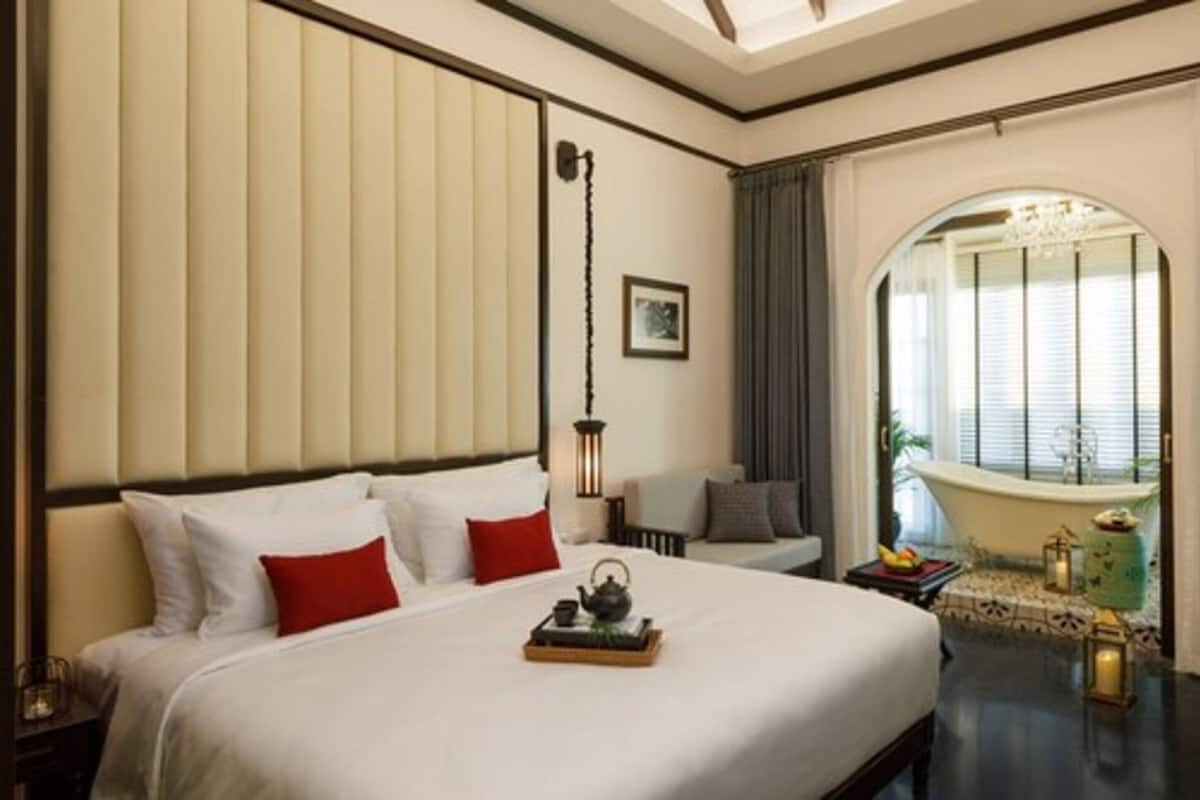 Habitación de hotel con una cama grande, una mesita de noche con una bandeja de té, una lámpara y una ventana con cortinas. Se ve una bañera a través de un arco.