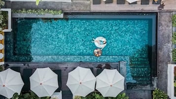 Una piscina rectangular con superficie de agua azul cristalina, rodeada de sombrillas blancas y abundante vegetación.
