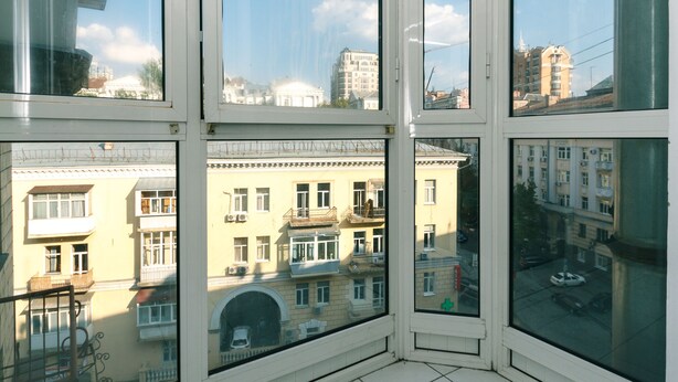 Una stanza d'angolo con ampie finestre, pavimento in piastrelle bianche e vista sugli edifici circostanti.