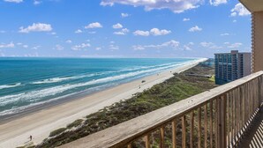 Une vue sur la plage depuis un balcon doté d’une balustrade en bois, avec l’océan en contrebas et un bâtiment en arrière-plan.
