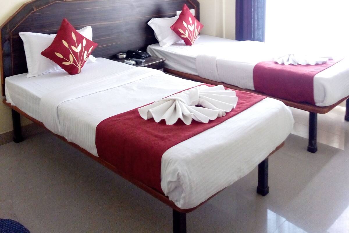 Habitación de hotel con dos camas, cada una con ropa de cama blanca y detalles rojos, un ventilador de techo y una ventana con cortinas.
