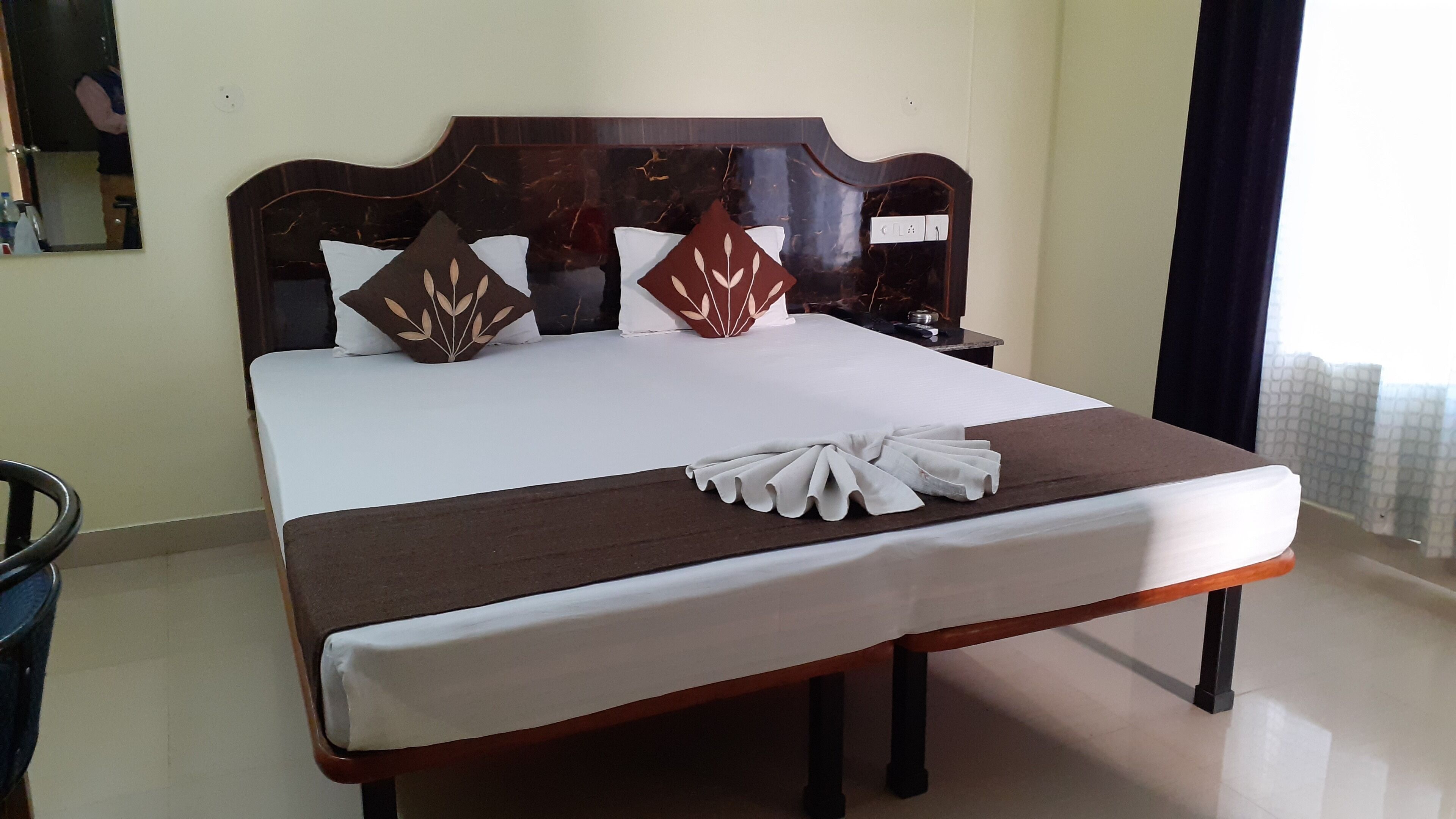 A neatly made bed with a brown headboard and white linens, a folded towel on the bed, and a nightstand with a lamp.