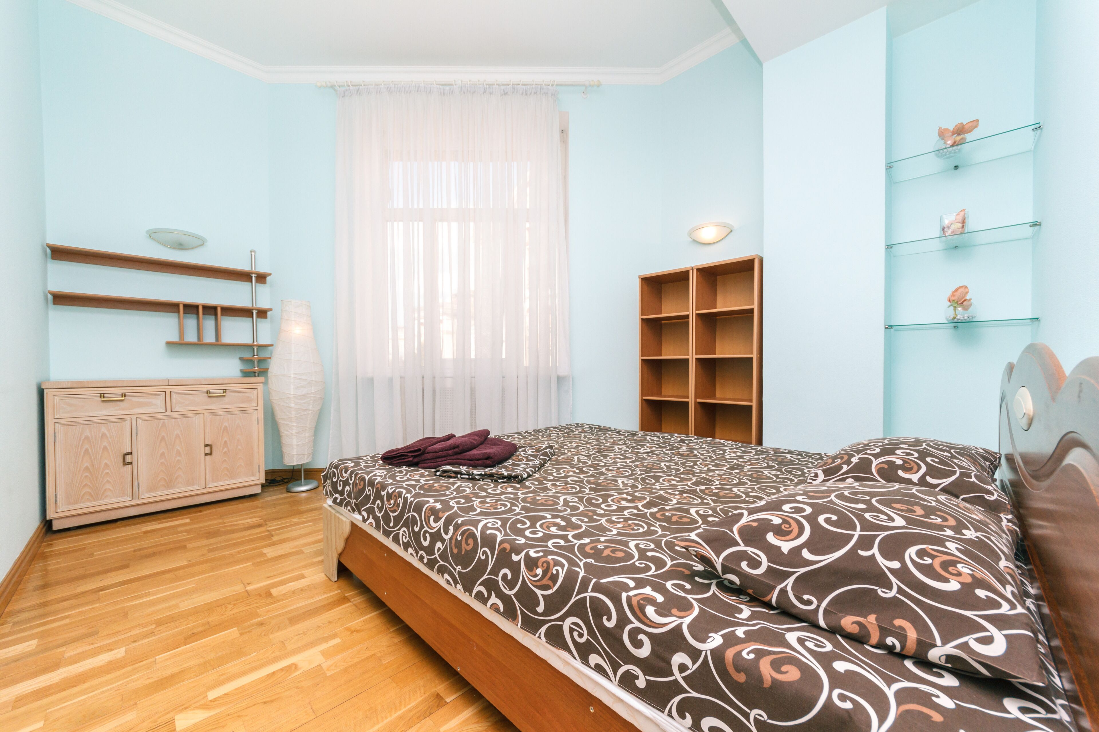 A bedroom with a wooden bed, a bedside table, a wardrobe, and a window with sheer curtains.