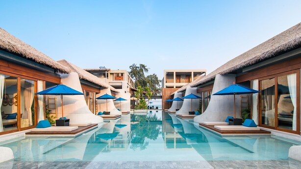 A pool area with thatched-roof buildings, blue umbrellas, and modern lounge seating. - Lullaby The Sea Hua Hin (Cha-am)