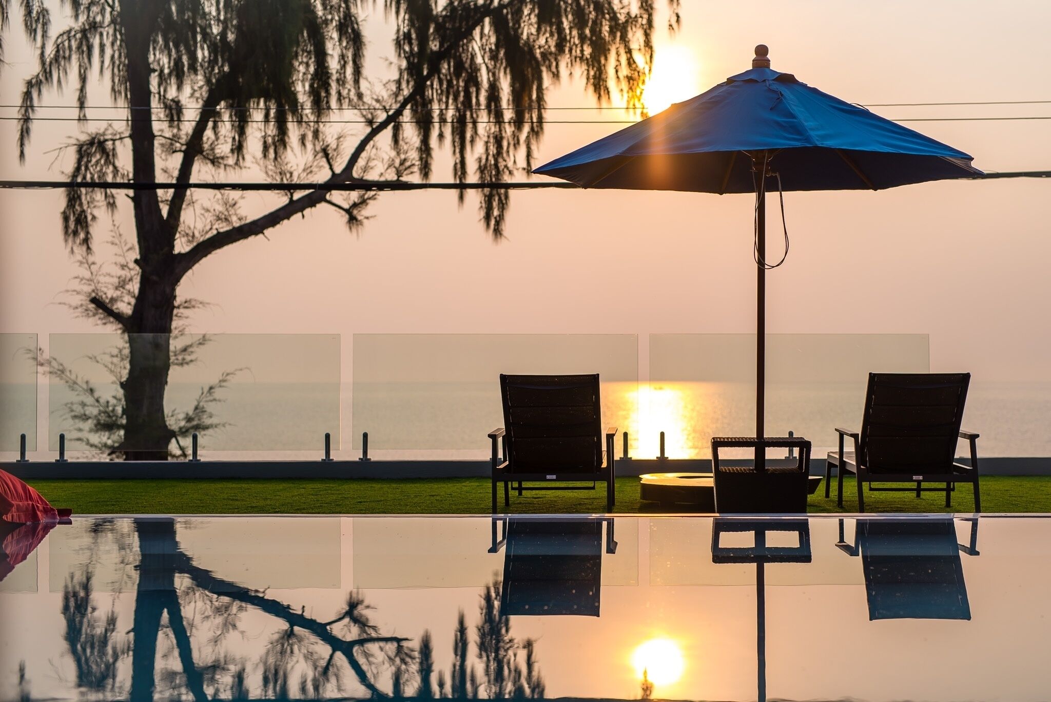 A poolside area with a blue umbrella, two lounge chairs, and a reflection on the water.