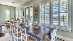 A dining area with a wooden table, white chairs, and large windows with shutters. - HEAVENLY VIEW: Spacious & Beautiful 30A Home with Pool, Huge Deck, Steps to Beach! (Santa Rosa Beach)