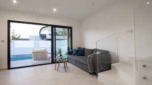 Una sala moderna con un sofá gris, un sillón blanco y una mesa de vidrio. Hay un ventanal corredero que da a una zona de piscina.