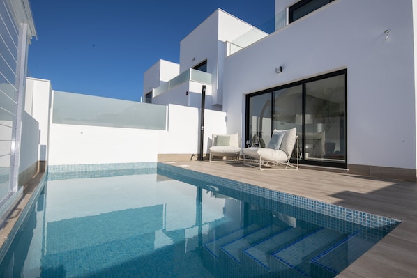 A modern outdoor swimming pool with clear blue water, surrounded by a wooden deck and white walls.