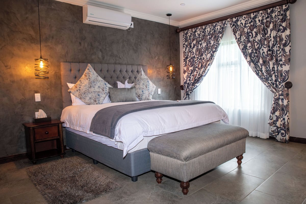 A bedroom with a tufted headboard, a grey upholstered bench, patterned curtains, and a textured rug.