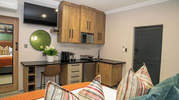 A compact hotel room with a kitchenette, a bed with striped pillows, a TV mounted above the counter, and a dark door.