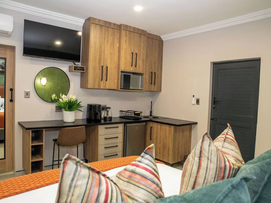 A compact hotel room with a kitchenette, a bed with striped pillows, a TV mounted above the counter, and a dark door.