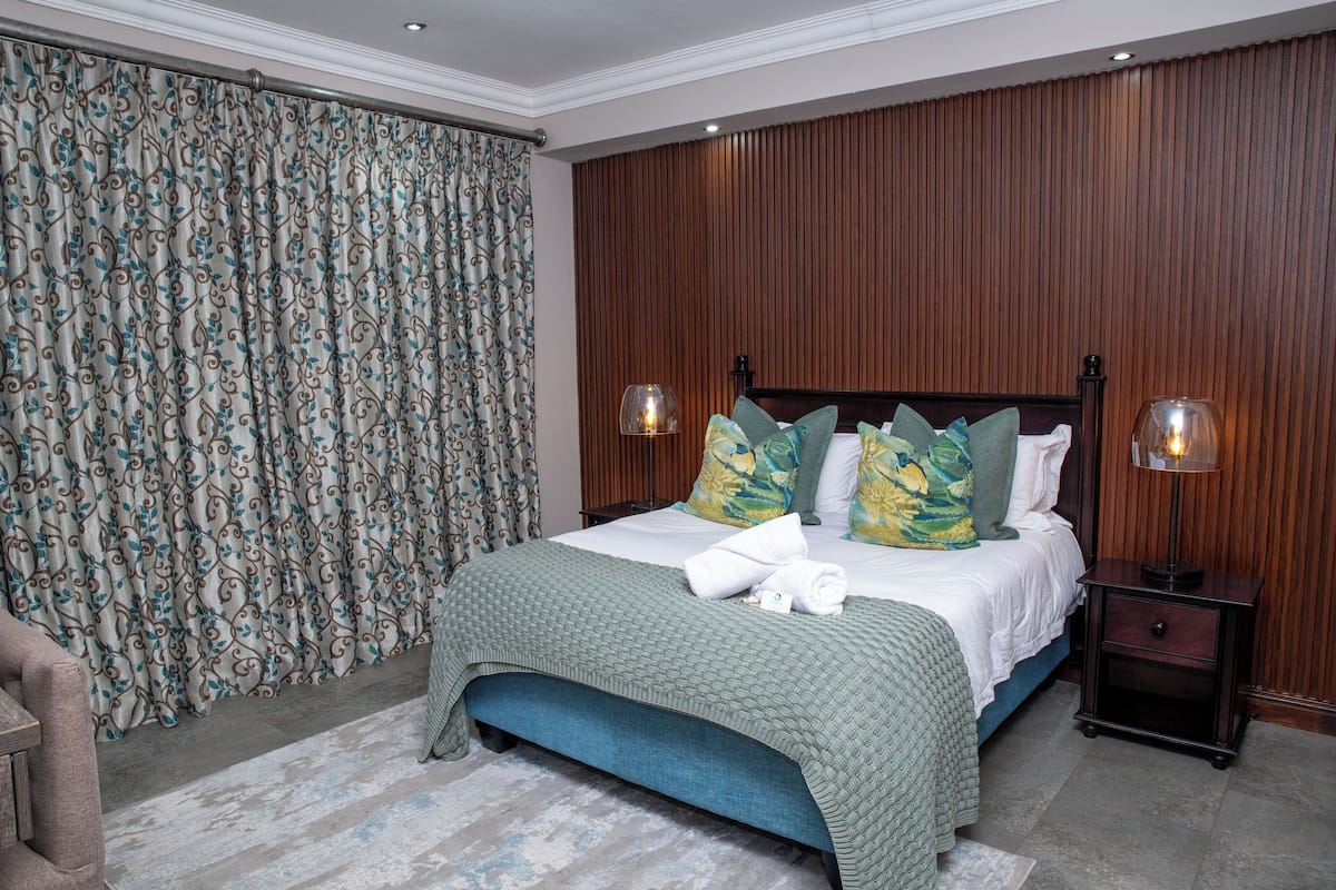 A neatly made bed with a green bedspread, two decorative pillows, and a matching green bench at the foot of the bed. There are two bedside tables with lamps, a wooden headboard, and a patterned curtain.