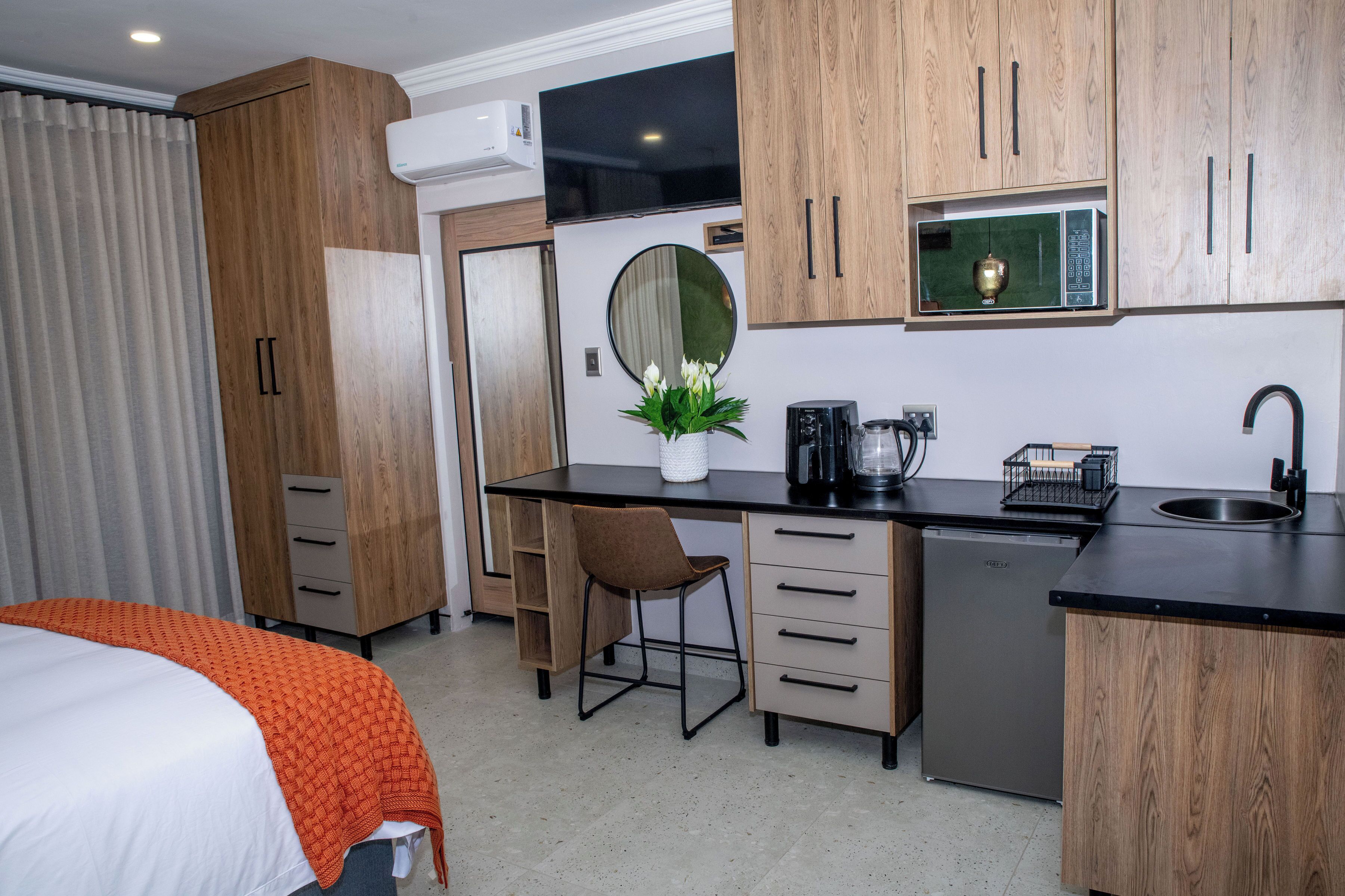 A compact hotel room with a bed, a desk with a chair, a small kitchen area with a microwave, and a sink.
