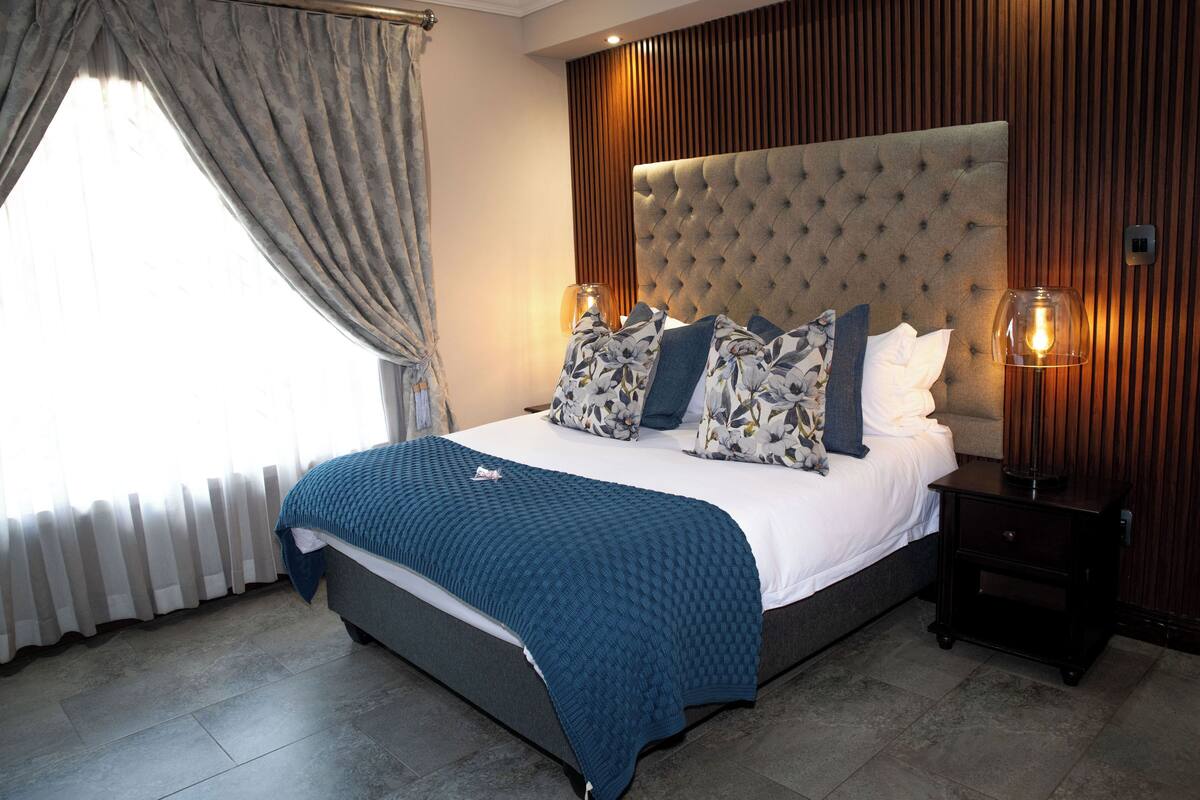 A neatly made bed with a blue blanket, a tufted headboard, and decorative pillows. There is a bedside table with a lamp, and the room has wooden paneling on the walls.