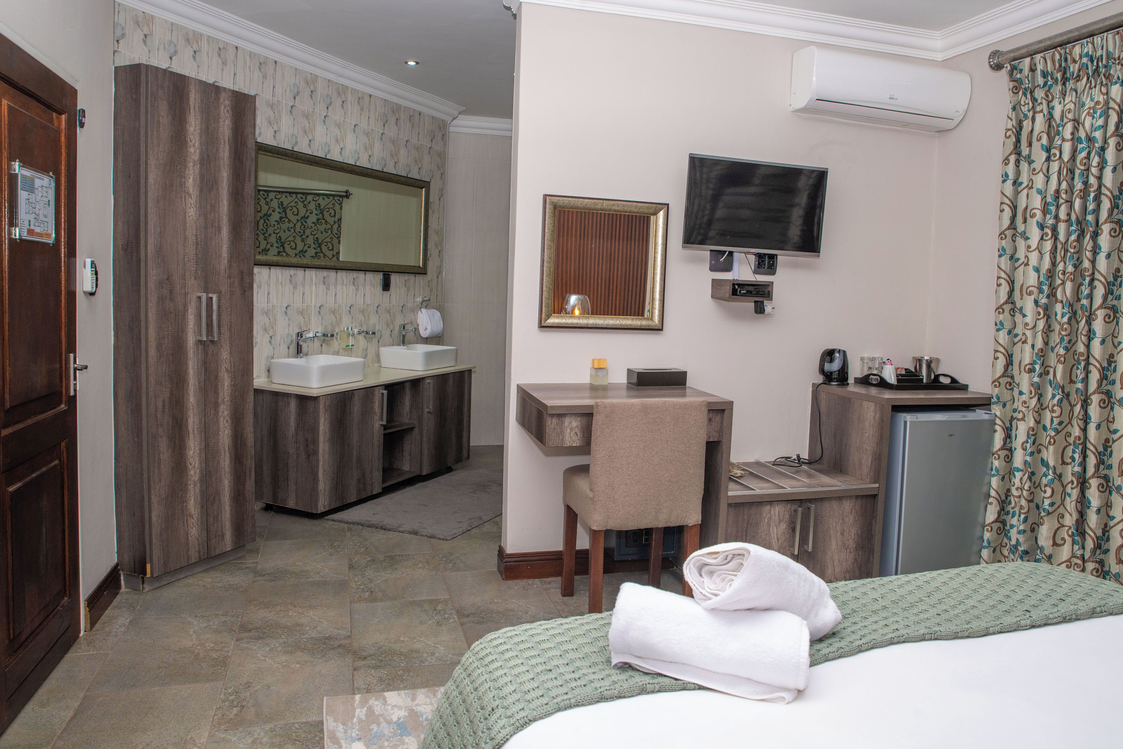 Habitación de hotel con cama, escritorio, silla, televisor, espejo, lavamanos y armario.