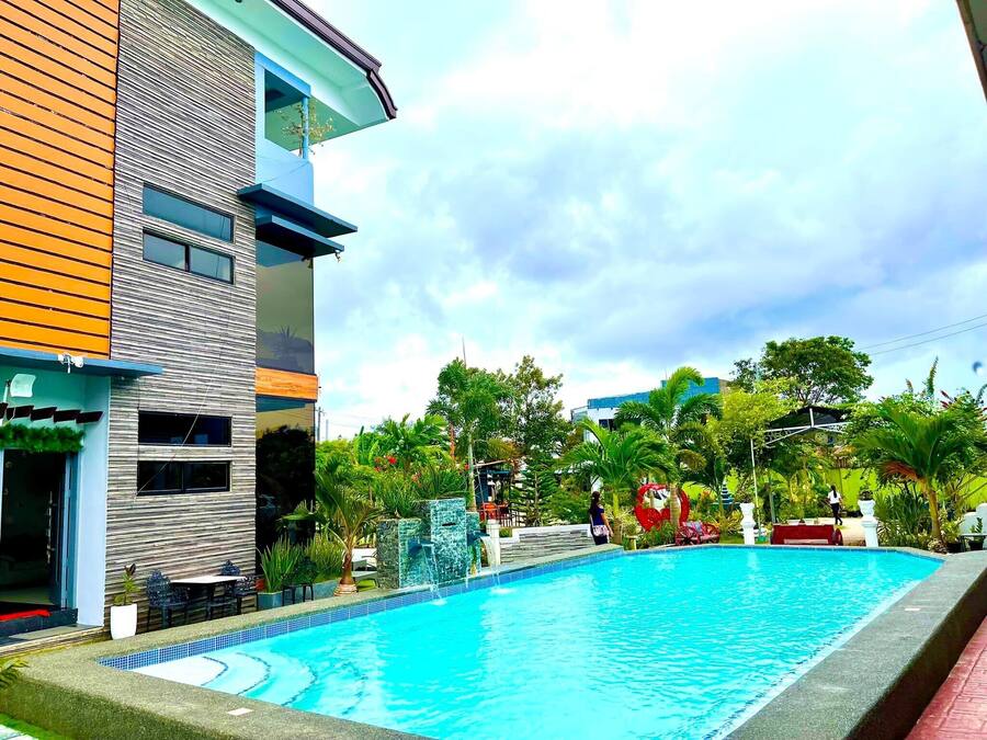 Un edificio moderno con piscina, mobiliario exterior y abundante vegetación.