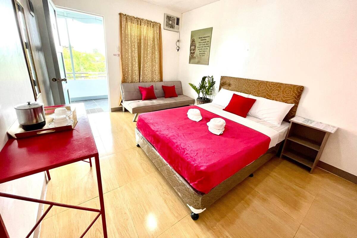 A hotel room with a bed, a sofa, a small table with a kettle, and a window with curtains.