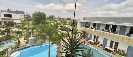 A view of a swimming pool with a large potted plant in the foreground, surrounded by buildings and greenery.