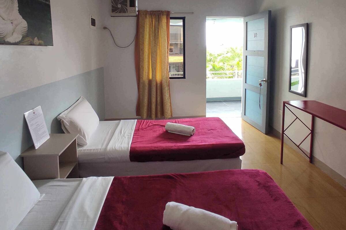 A hotel room with two beds, a window with curtains, an air conditioning unit, a door to a balcony, and a small table.