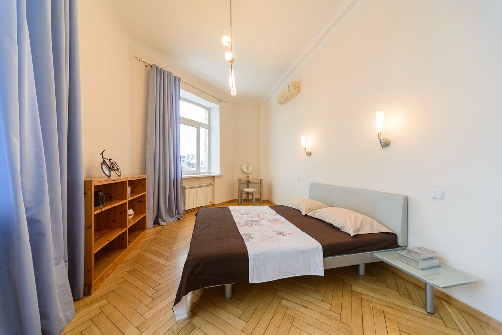A bedroom with a bed, a small bedside table, a wooden shelf unit, a window with curtains, and a city view.