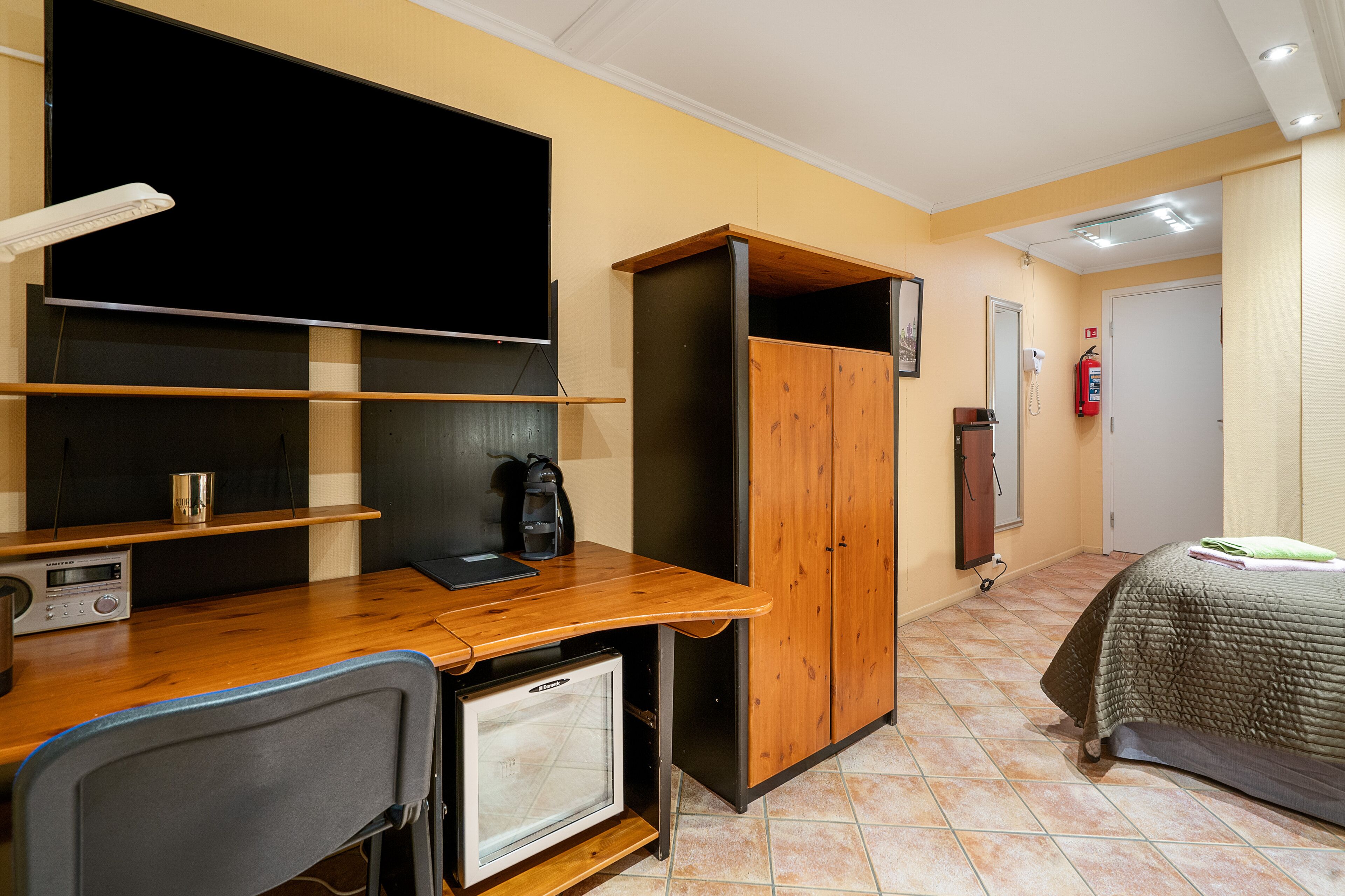 A compact room with a wooden desk, a small refrigerator, a bed with a green blanket, a television, and a fire extinguisher on the wall.