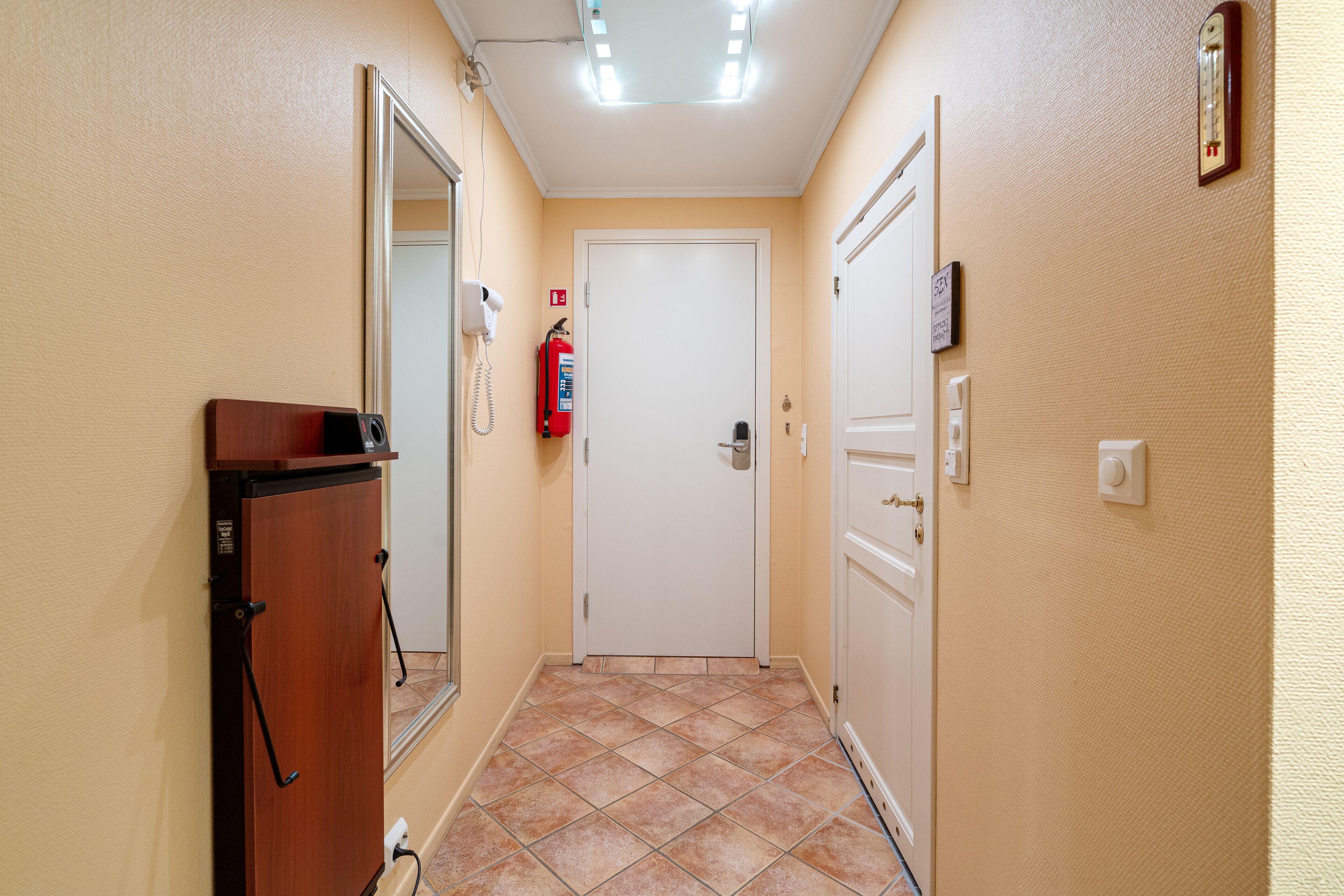 A narrow hallway with a wooden cabinet, a white door, a fire extinguisher, and a thermostat on the wall.