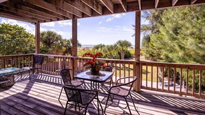 Una terraza de madera con una mesa y sillas de vidrio, con vistas a un paisaje de árboles y un cuerpo de agua.