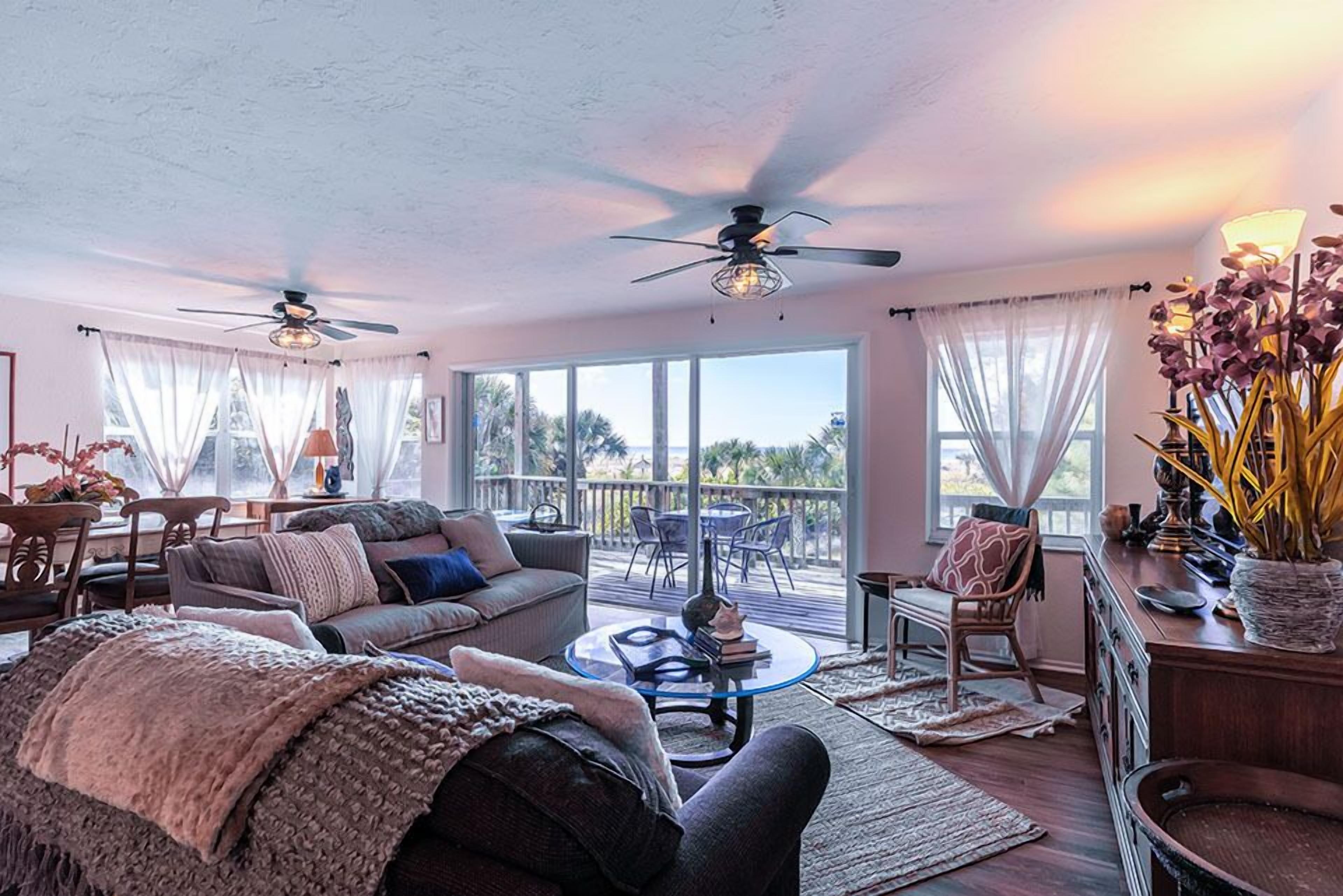 A spacious living room with a sofa, coffee table, and chairs, large windows with curtains, ceiling fans, and a view of palm trees.