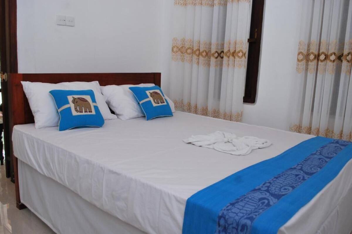 A neatly made bed with white bedding and two blue pillows with elephant designs, a blue and white patterned bedspread, and white curtains with a gold pattern.