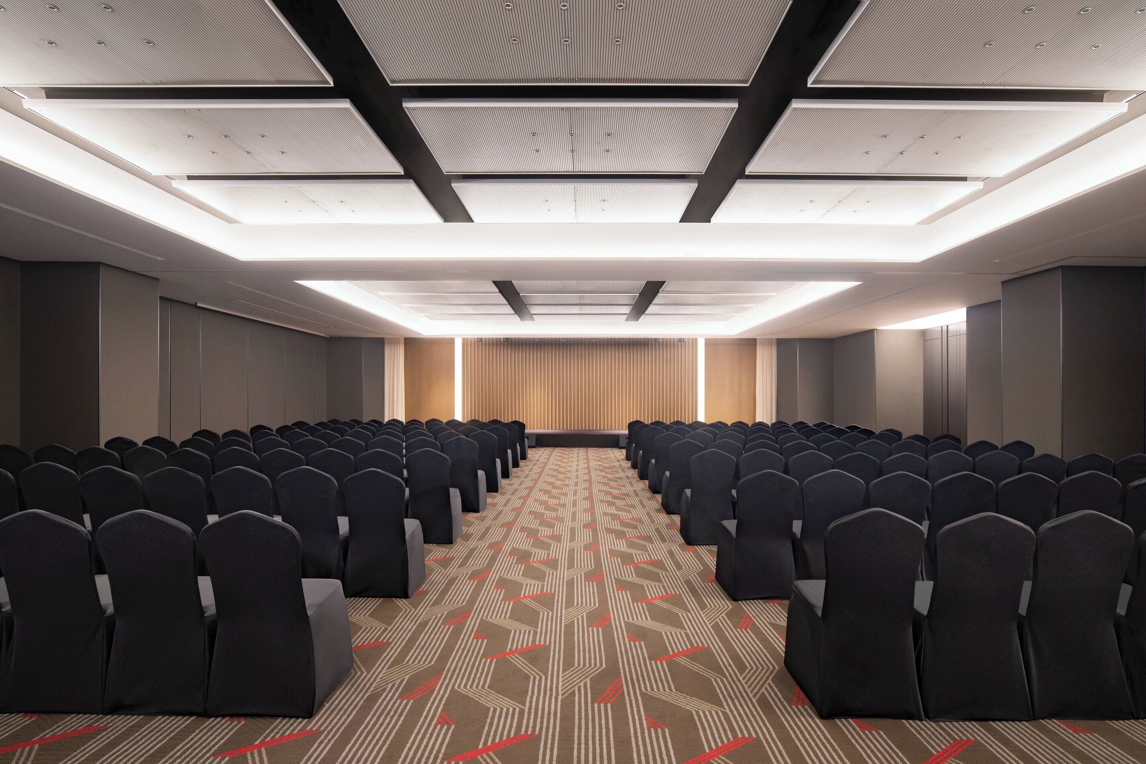 A conference room with rows of chairs, a patterned carpet, and a ceiling with recessed lighting.