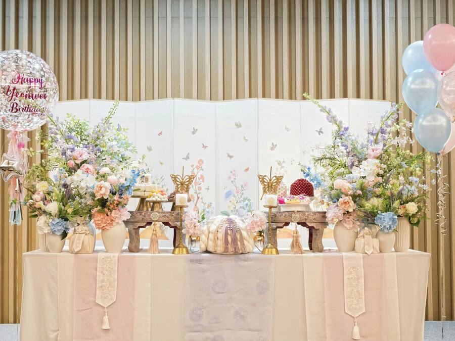 A decorated table with floral arrangements, a 'Happy Birthday' balloon, and a backdrop with vertical lines.