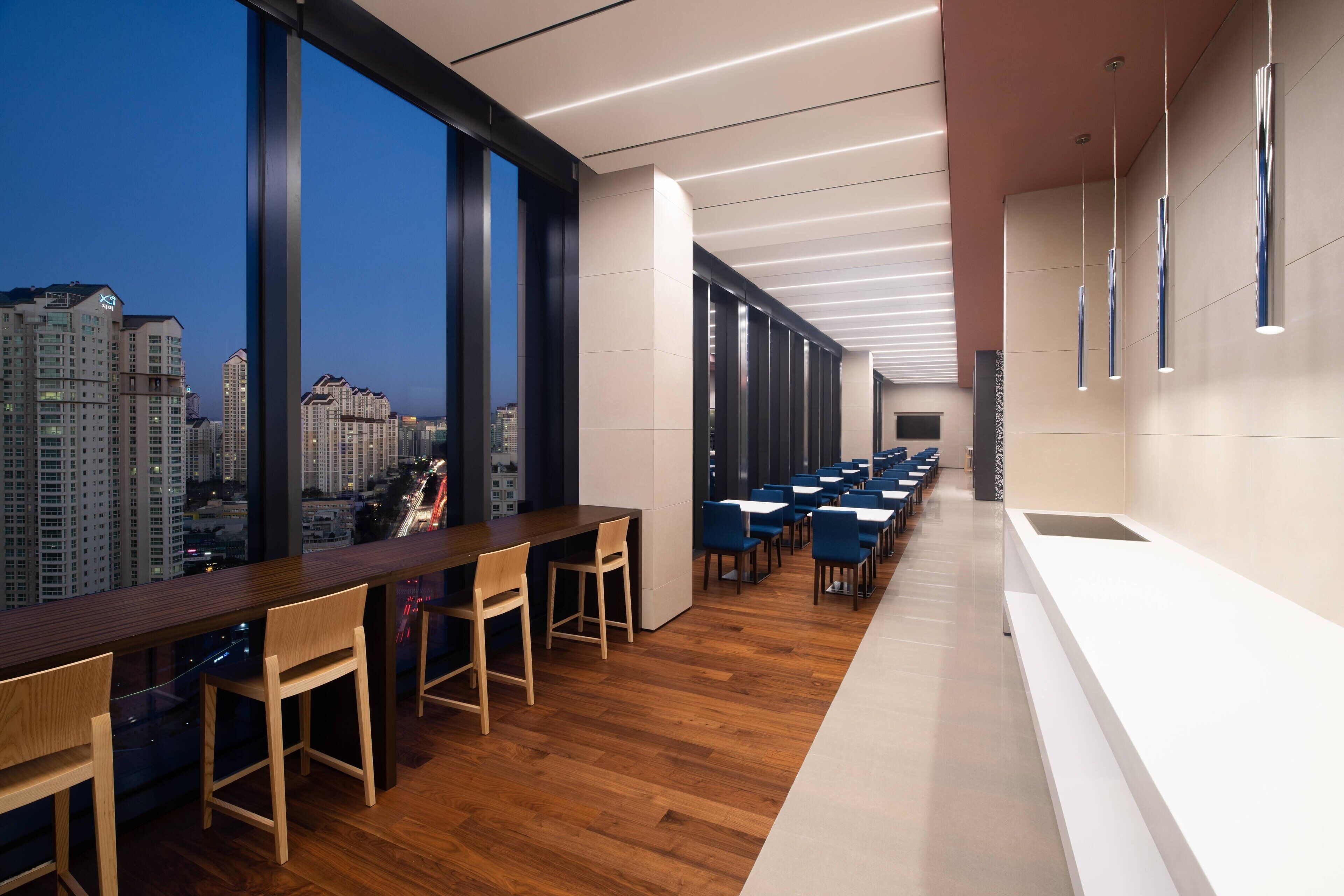 A modern interior with a long counter and chairs, large windows offering a city view, and a long row of tables.