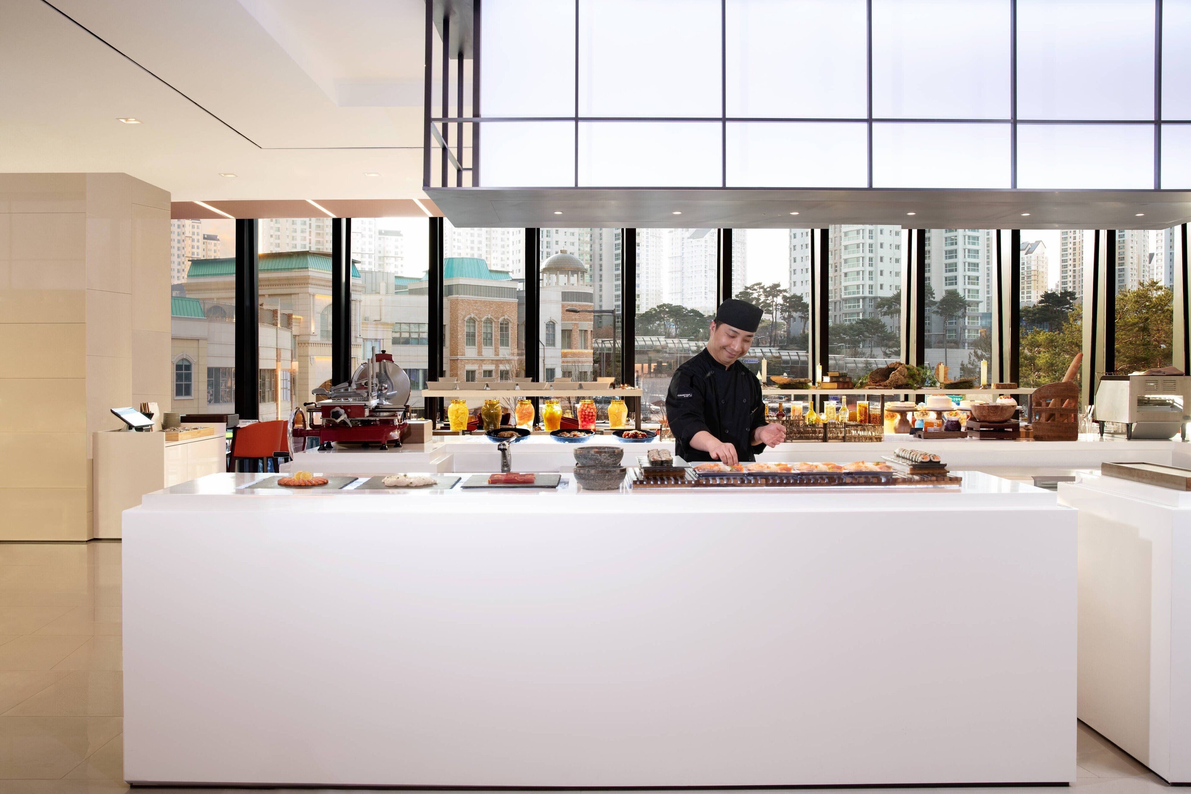 A modern breakfast buffet with a chef preparing food, large windows offering city views, and a clean, contemporary interior design.