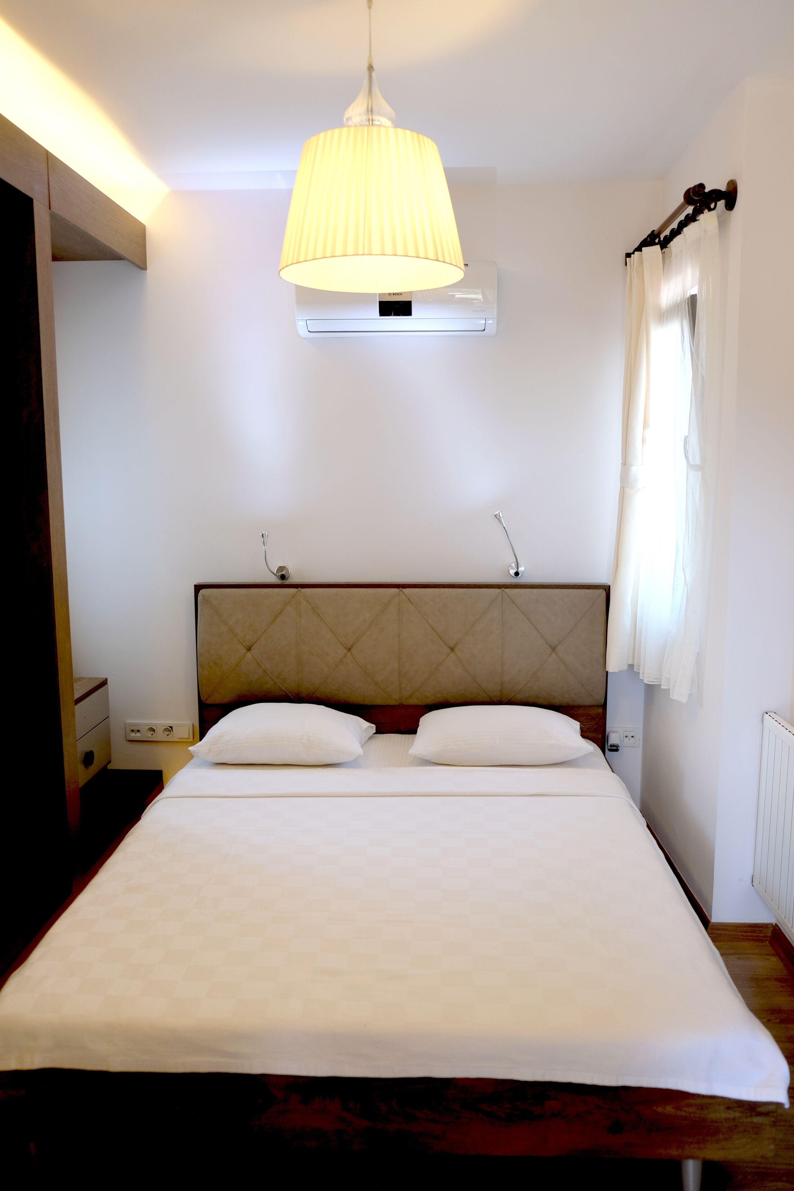 A neatly made bed with a headboard, a bedside lamp, and a window with curtains.