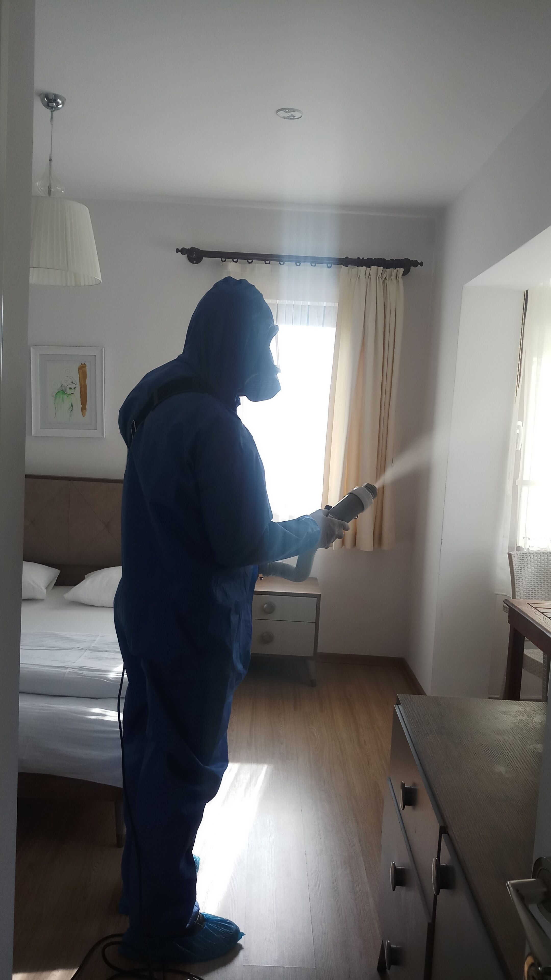 A person in a blue protective suit using a spray device in a hotel room with a bed, a nightstand, and a window with curtains.