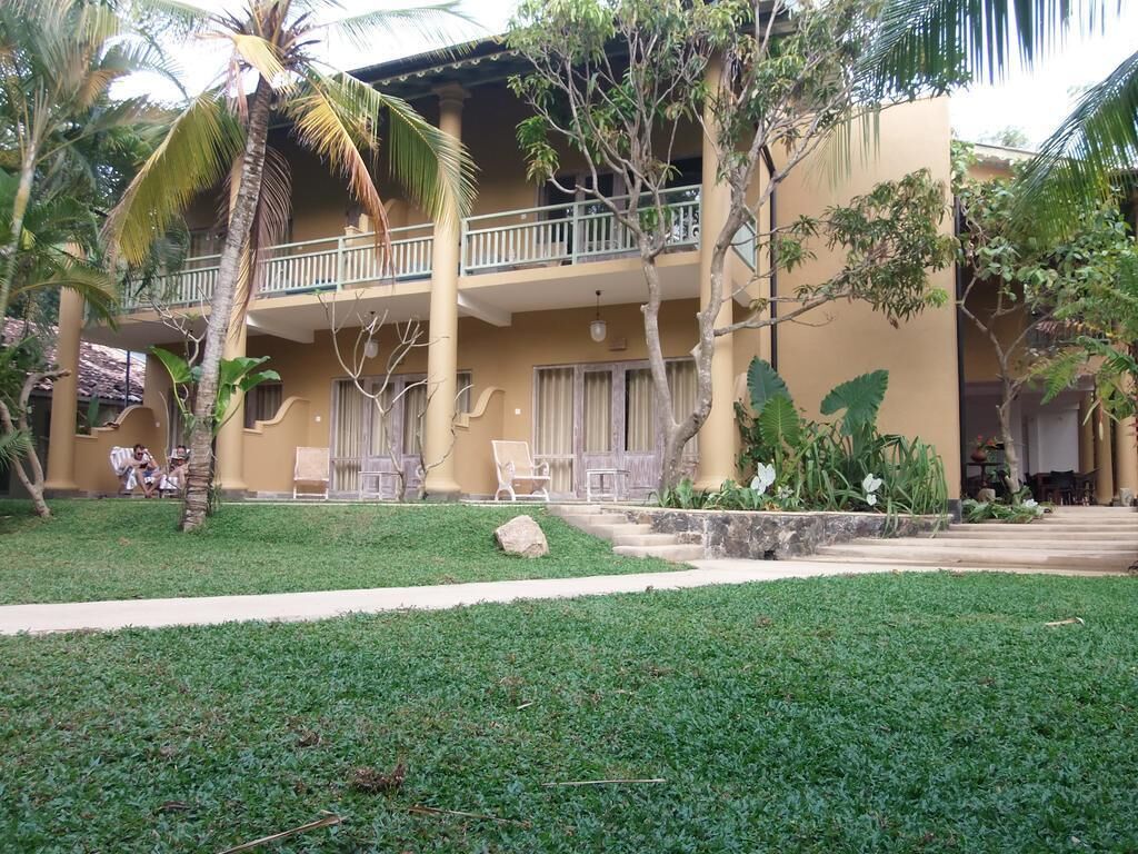 A two-story building with a balcony, surrounded by greenery and palm trees.