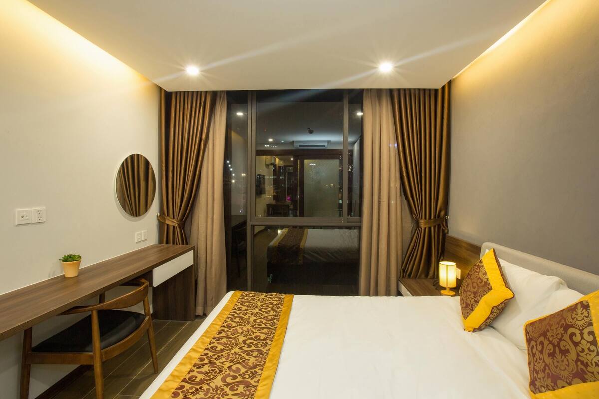 A hotel room with a bed, a desk, a chair, a nightstand with a lamp, and a view of the city through large windows.