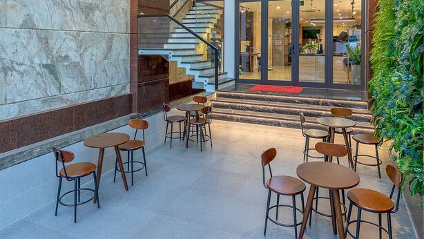 A lobby with a marble wall, wooden ceiling, and a staircase leading to an upper level. There are tables and chairs arranged for seating. - Citypoint Apartment (Da Nang)