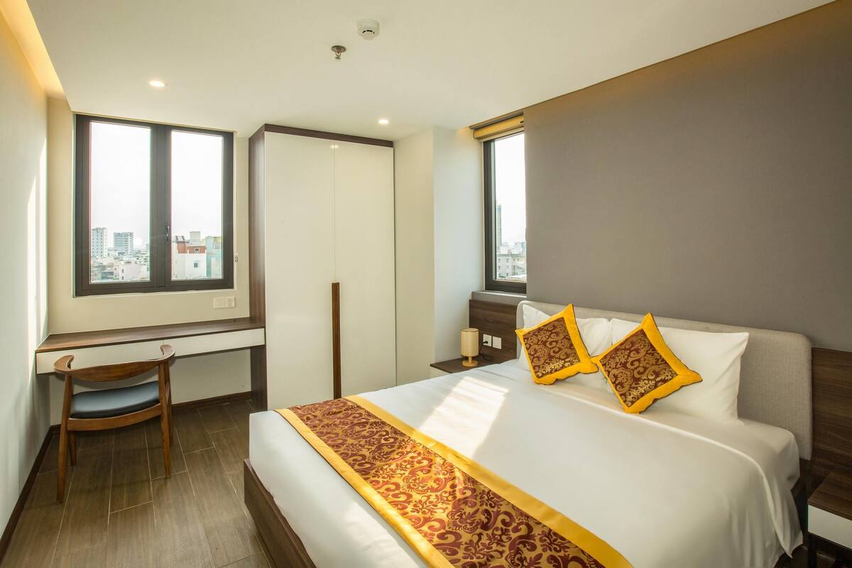 A hotel room with a large bed, a desk, and a chair. There are two windows providing a view of the city.