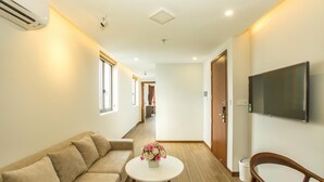 A hotel room with a sofa, a small round table with a flower vase, a TV mounted on the wall, and a wooden chair. - Citypoint Apartment (Da Nang)