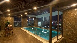 A modern indoor pool area with a city view, a wooden chair, and a vase of flowers. - Citypoint Apartment (Da Nang)