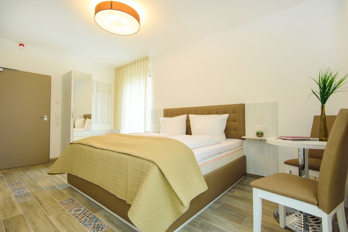 A hotel room with a bed, a chair, a table, and a plant.